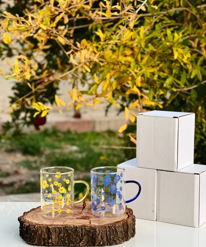 لیوان پیرکس دستهدار طرح گل بابونه زرد و آبی روی زیرلیوانی چوبی